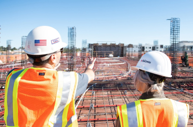Two construction workers in safety vests and helmets stand at a building site, with one pointing toward the construction area filled with rebar and materials. Buildings and a clear sky are visible in the background.