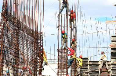 Construction workers wearing safety gear climb and work on tall metal scaffolding and rebar structures at a building site under a partly cloudy sky.