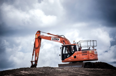 An orange excavator labeled "Lynch" sits on top of a mound of dirt under a cloudy sky, appearing to dig or move soil at a construction or earthmoving site.