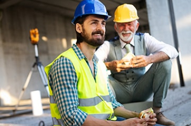 Two male construction workers wearing safety vests and hard hats sit and enjoy a lunch break, smiling and relaxed, at a construction site. One holds a sandwich, and measuring equipment is visible in the background.