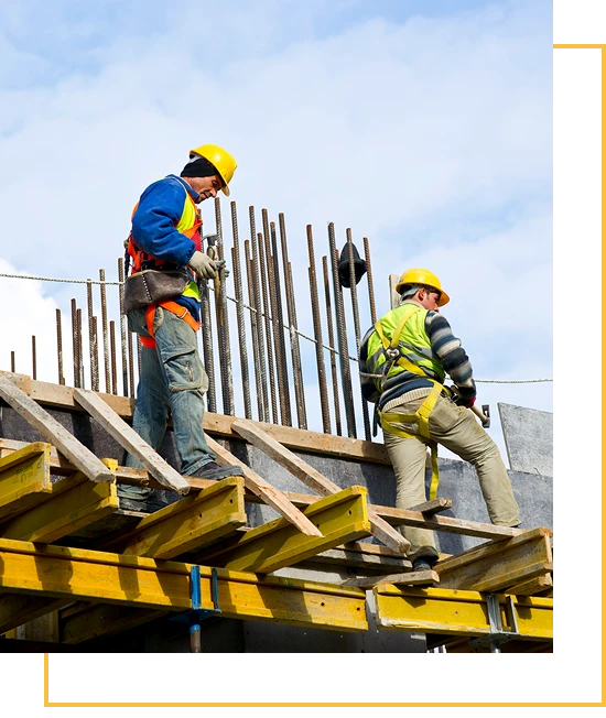 Two construction workers in safety gear and helmets work on a building framework, standing on wooden beams with metal rods in the background under a partly cloudy sky.
