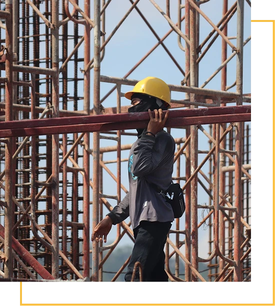 A construction worker wearing a yellow hard hat and face covering carries a metal beam on his shoulder, walking past scaffolding and metal bars at a construction site.