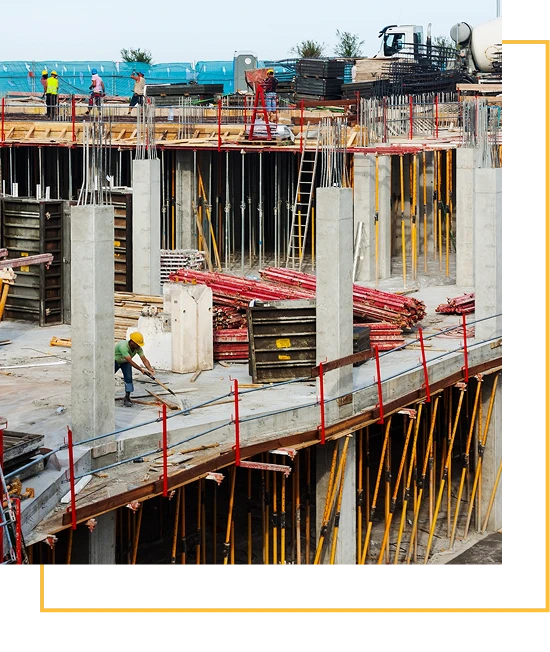 Construction site with workers, concrete columns, scaffolding, ladders, and various building materials. Several workers are wearing safety helmets and vests while working on the structure’s upper level.