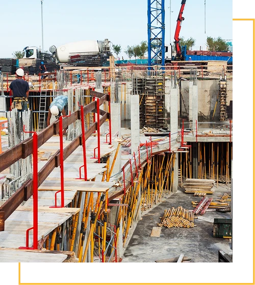 A construction site with workers, concrete columns, scaffolding, and building materials. A cement mixer truck and cranes are visible in the background, with safety railings along the unfinished structure.