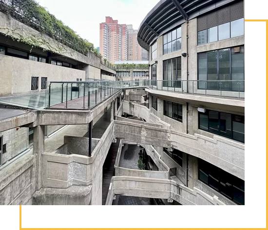 A multi-level concrete building with curved walkways and glass railings, featuring modern architecture. In the background, tall apartment buildings are visible under a cloudy sky.
