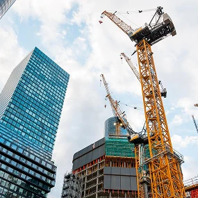 Several tall construction cranes operate among modern glass skyscrapers and unfinished buildings against a partly cloudy sky, illustrating ongoing urban development in a cityscape.