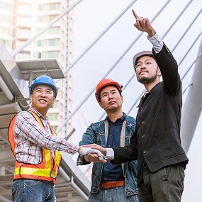 Three men, two in safety helmets and vests and one in business attire, stand outdoors near a modern structure. Two shake hands while the third points upward, suggesting discussion or planning at a construction site.