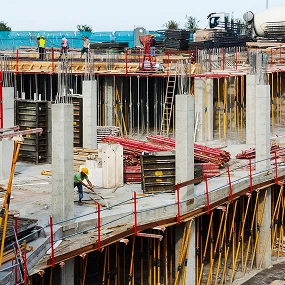 Several construction workers are working on a large multi-level building site with concrete columns, scaffolding, metal bars, and construction materials scattered around.