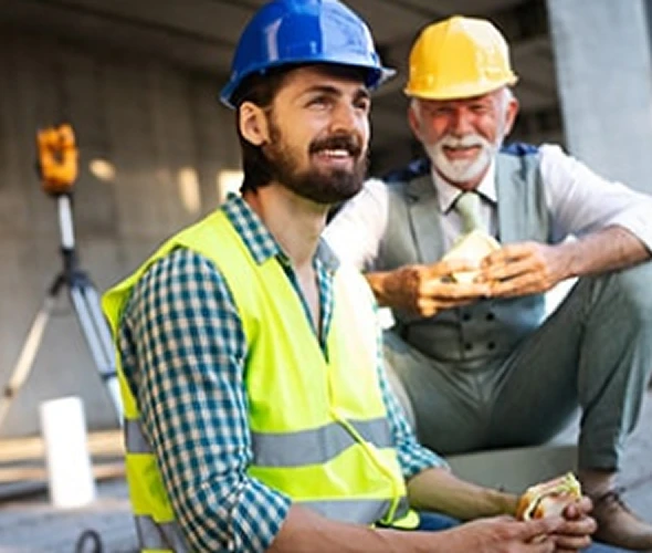 Two construction workers wearing safety vests and hard hats are smiling and taking a break, sitting outdoors at a worksite while holding sandwiches.