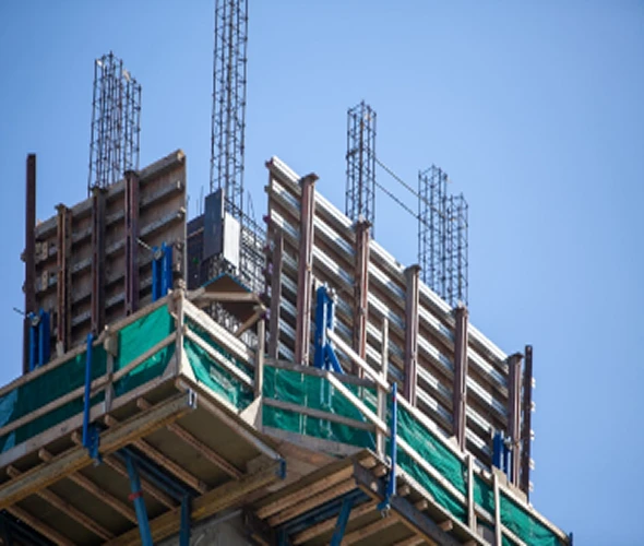 A close-up view of a building under construction, showing scaffolding, wooden planks, and steel reinforcement bars extending upwards against a clear blue sky.