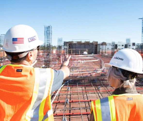 Two construction workers wearing hard hats and reflective vests stand on a building site, one pointing toward the unfinished structure in front of them under a clear blue sky.