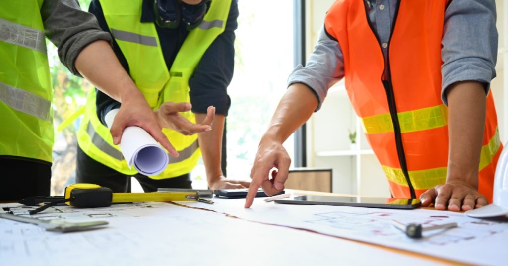 Two people in safety vests review blueprints on a table, one holding rolled plans and the other pointing at the documents, with measuring tools and a tablet also visible.