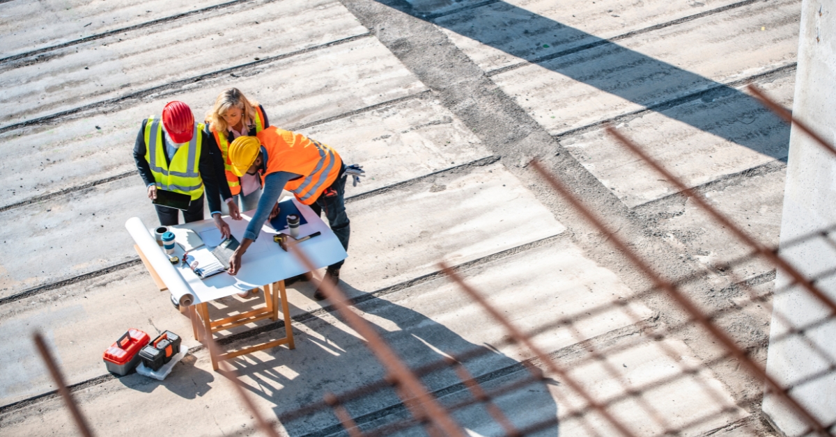 Three construction workers in safety vests and helmets review blueprints on a table at an outdoor work site, viewed from above. Construction tools and materials are nearby.