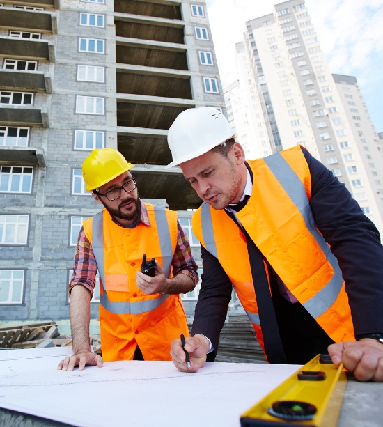 Two construction workers in safety vests and hard hats review blueprints at a construction site, with a partially built high-rise building and cityscape in the background. One man writes while the other holds a walkie-talkie.